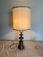 Front view of the tall classic lamp with off-white cylindrical fabric shade and bronze turned base, placed against wall with power cord visible.