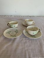 Three teacups and three saucers on a table. Two sets have green clover painted design and textured surface with brown handles. One set is white with a ribbed texture.
