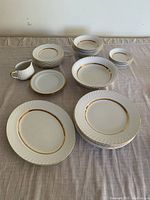 Wide view of the dishware set on table, showing platter, plates, bowls, saucers and small milk jug all white with gold trim.