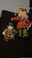 Photo showing two decorative seasonal scarecrow figurines on a dark couch.