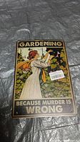 Photo of rectangular metal sign with vintage-style illustration of woman gardening and text 'Gardening Because Murder Is Wrong'.