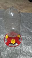 Photo showing a clear plastic hummingbird feeder bottle with a red plastic base and four yellow flower-shaped feeding ports. The feeder is placed on a gray tarp on the floor.