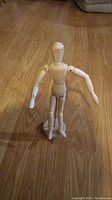 Photo of standing wooden articulated mannequin figure on hardwood floor, showing movable arm and leg joints and attached wooden base.