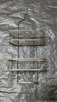 Photo of metal two-tier shower caddy hanger with hook for bathroom storage, showing its full front view against a gray background.