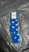 Blue socks with white smiley faces pattern by Super H, laid flat on grey surface with packaging tag attached.