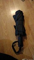 Photo of a folded black umbrella lying on a wooden floor, showing the handle and wrist strap.