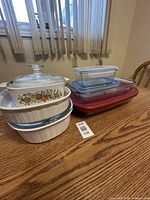 Multiple glass and ceramic containers including two Anchor food storage containers and three casserole dishes on wood table.