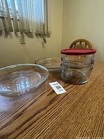 Photo showing two clear glass pie plates, one shallow and one deeper, and three clear round glass storage containers stacked with red lids on a wooden table.