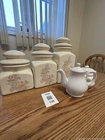 Photo of three ceramic canisters with floral design and a white ceramic teapot on a wooden table under natural light, showing all items together.