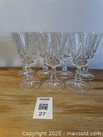 Photo showing six crystal wine glasses with detailed etched pattern on bowls, faceted stems, on wooden surface against plain background.