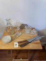 Overview of glass decanter, crystal candle holder, small pitcher, lidded dish, decorative metal piece, three decanter stoppers, four crystal bowls, two knives with wooden handles on wooden table