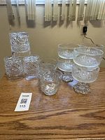 Photo showing set of six crystal cut glass rocks glasses stacked and four parfait bowls with decorative bases arranged on a wooden surface under window blinds.