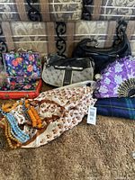 View of two Guess handbags, jewelry box, multiple necklaces, scarf, purple clutch, and tartan cloth arranged on a carpeted area with a patterned sofa in background