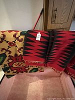 Two handwoven rugs shown draped over a piece of furniture. One is red and black zigzag pattern and the other is multicolor geometric pattern.