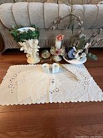 Full lot arranged on couch seat including white embroidered linens, porcelain cherub candlestick with artificial greenery, multiple floral decorative pieces, small clock, and gold-tone trinket.