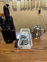 Kitchen knives set in block, white basket with wine accessories, and stainless steel teapot on wooden table under window curtains