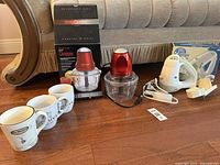 Full lot photo showing Betty Crocker food chopper box and unit, Ergo hand mixer with attachments and cords, and four ceramic mugs with printed designs.