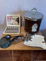 Photo showing Mikasa spoons in box, vintage ice bucket, stone coasters, silver napkin holder, and decorative plates on wooden table.