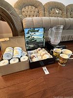 Full lot of miscellaneous mugs, teacups, and a glass pitcher arranged on floor in front of couch
