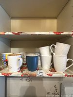Shelf with multiple ceramic mugs of varying designs and sizes, including white textured mugs, solid blue, and colorful printed mugs.