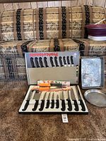 Photo of closed and open knife case showing eleven knives with black handles and extra yellow handle set in packaging, displayed on patterned sofa.