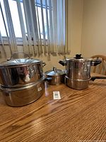 Five stainless steel pots of various sizes on wooden table under window with vertical blinds. Pots have glass lids and black handles. No brand markings visible.