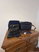 Four bags displayed on a wooden dresser: two black leather purses and two dark blue fabric bags with straps.