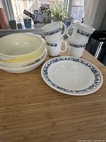 Photo showing 4 white Corningware mugs with blue floral pattern stacked, 2 yellow Pyrex bowls with floral designs, and 1 Corelle plate with matching blue floral border on wooden table