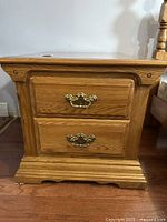 Front view of one wooden side table showing two drawers with brass handles and carved detailing on base.
