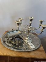 Photo showing the five-arm silver plated candelabra, large round serving tray, rectangular serving dish, and decorative napkin or letter holder all placed on a wooden surface.