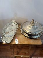 Overview of the silver plated serving dishes on a wooden surface, showing oval dish with glass insert and paired tongs, alongside round covered serving dish.