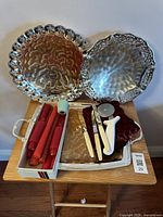 Photo showing two silver trays, red candles, white candle holder, rectangular hammered tray, pizza cutter, butter knife, and red cloth on a small wooden table.
