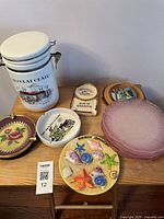 Photo showing white ceramic canister labelled 'Chocolat Chaud', multiple small decorative ashtrays, pink ribbed glass plates, and a colorful sea creature themed decorative plate.