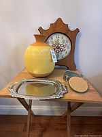 Front view of vintage decor items: yellow ceramic vase, wooden floral clock, silver-tone tray, and coasters on a small wooden table.