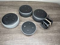 Photo of four Amazon Echo Dot smart speakers on a wooden surface, showing three 4th generation dark grey spherical units and one smaller 3rd generation light grey unit with a flat top, alongside one power cord.