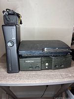 Photo showing Xbox 360, PlayStation 3 Slim and Original Xbox consoles stacked on a wooden surface.