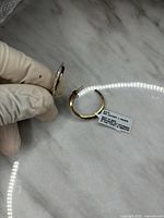 Two images showing gold-tone hoop earrings with diamond accents held with gloved fingers, placed on a marble surface. A tag on earrings labeled 'DIA ACCENT J HOOPS'.