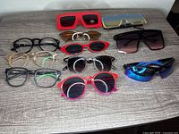 Lot of 11 sunglasses and eyeglasses on grey wooden surface, various frame and lens colors visible.