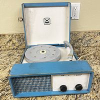 Front and interior view of blue and white portable record player phonograph with tonearm, turntable, speaker grille, and control knobs.
