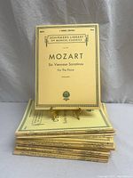 Stack of 20 yellow-covered antique music sheets with Mozart sheet displayed upright on a stand, showing title 'Mozart Six Viennese Sonatinas for the Piano'.