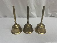 Front view of three vintage brass bells side by side, showing their tapered handles and engraved bodies.
