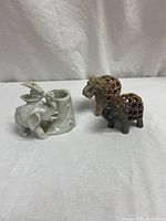 Photo showing the set of four figurines: a light gray soapstone mother elephant with baby in hollow cavity attached to a cylindrical base, two brown soapstone elephants with carved latticework, and one carved jade elephant with small chip on ear.