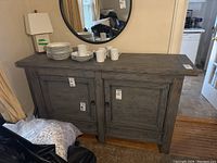 Front view of the two-door cabinet with plates and mugs on top, round mirror behind.