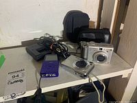 Lot overview showing digital cameras, charger with cord, camera carrying cases, lot identification tag