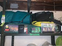 Wide view showing all camping gear including sleeping bag, arm chair in green carrying case, ponchos, hatchet, matches, and small light on a shelf.