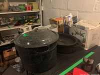 Large black speckled canning pot with lid on black surface, surrounded by other boxed kitchen items in basement setting