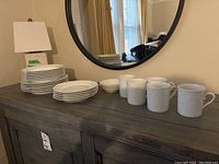 Dresser surface showing stacks of plates, bowls, and mugs in white ceramic with embossed botanical designs.