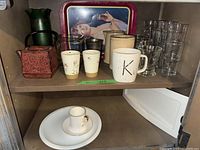 Two shelves of dishes including assorted cups, plates, mugs, glasses, Royal Albert teacup and saucer, cast iron teapots and a decorative tray.