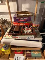 Overall view of stacked board games and game sets including Backgammon set, Balderdash, Chess & Checkers, Monopoly, a tin container with game pieces, a rolled-up poster or mat, and smaller boxed games beneath.