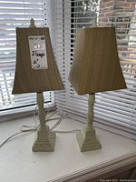 Pair of matching table lamps with cream composite bases and beige rectangular fabric shades, sitting on white window ledge with blinds in the background.